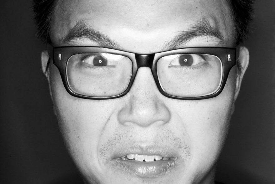 Close up of the face of a person wearing glasses, with raised eyebrows, their mouth in a slight sneer; black and white image.