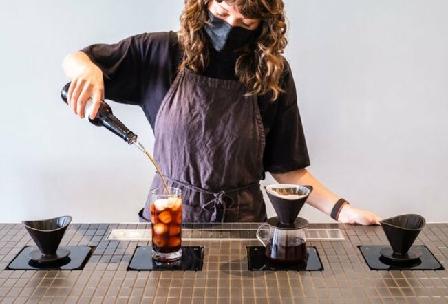 Barista with curly brown hair in a grey apron pouring cold brew coffee into a glass with lychee next to a slow drip coffee.