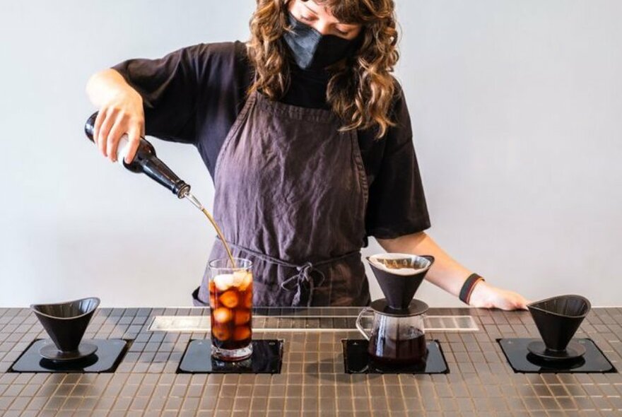 Barista with curly brown hair in a grey apron pouring cold brew coffee into a glass with lychee next to a slow drip coffee.