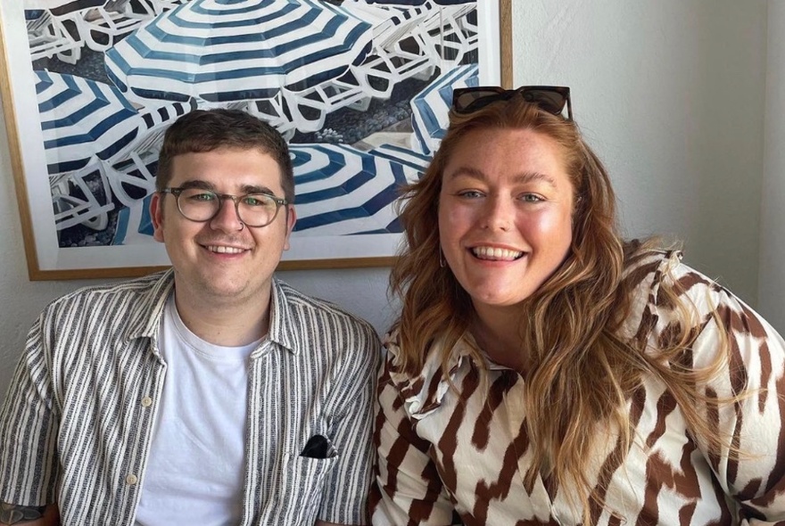 Two smiling people, seated in front of an artwork.