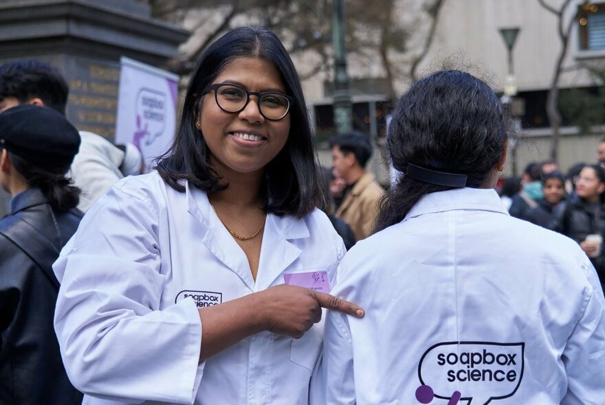 Two people in white science lab coats, standing outdoors in a public space, one with her back to the viewer and a stitched sign on her lab coat which reads 'soapbox science', the person facing the viewer smiling and pointing at the message.