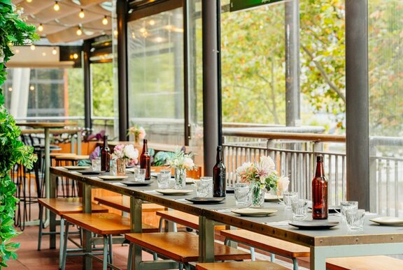Long tables set for lunch with bench seats in a restaurant with a leafy outlook.