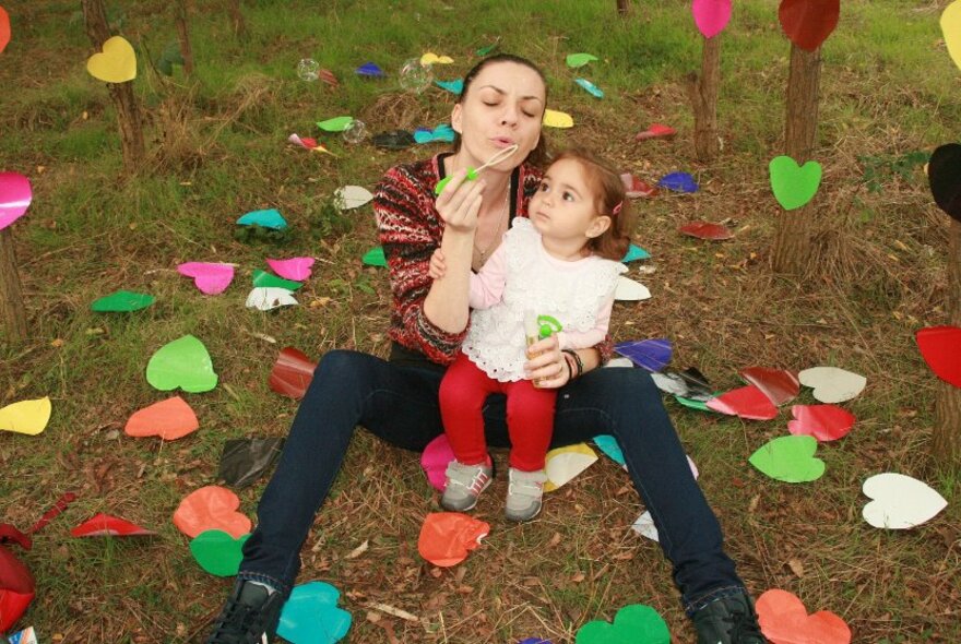 A woman sits on grass strewn with coloured paper hearts, a child seated on her lap. 