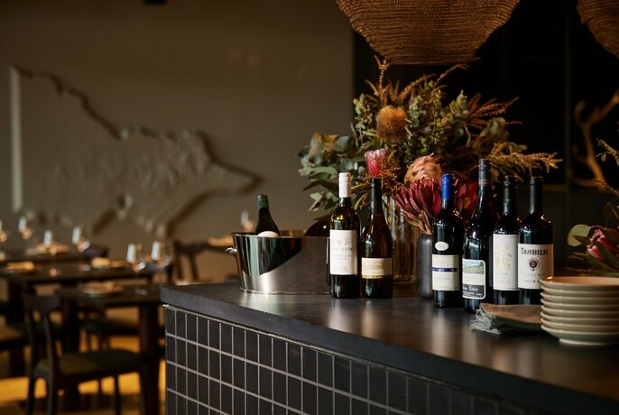 Interior of restaurant showing bottles of wine and and a floral arrangement on a dark counter, with a table set for service in the background.