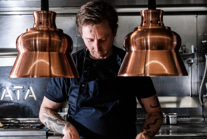 Chef Stephen Clark working at a grill under suspended copper lights.