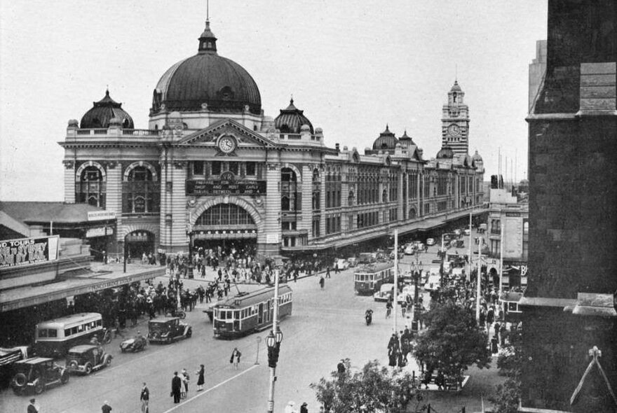 Black and white photo of Flinders Street Station with trams, cars and lots of pedestrians