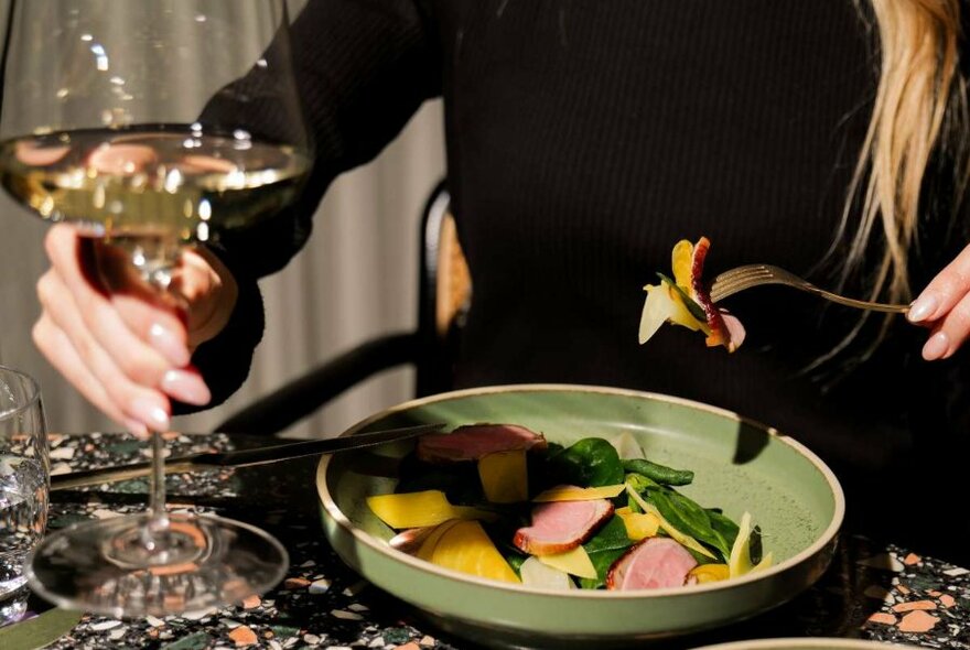 A person's hand raising a fork of food from a plate and holding a wine glass in their other hand