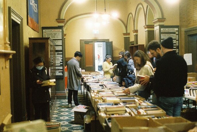 People browsing tables of books in a Victorian-era basement with decorative arches.