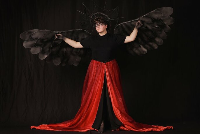 Person standing in a long red skirt with a train, black top and wearing black outstretched wings behind her; posed against a black background.