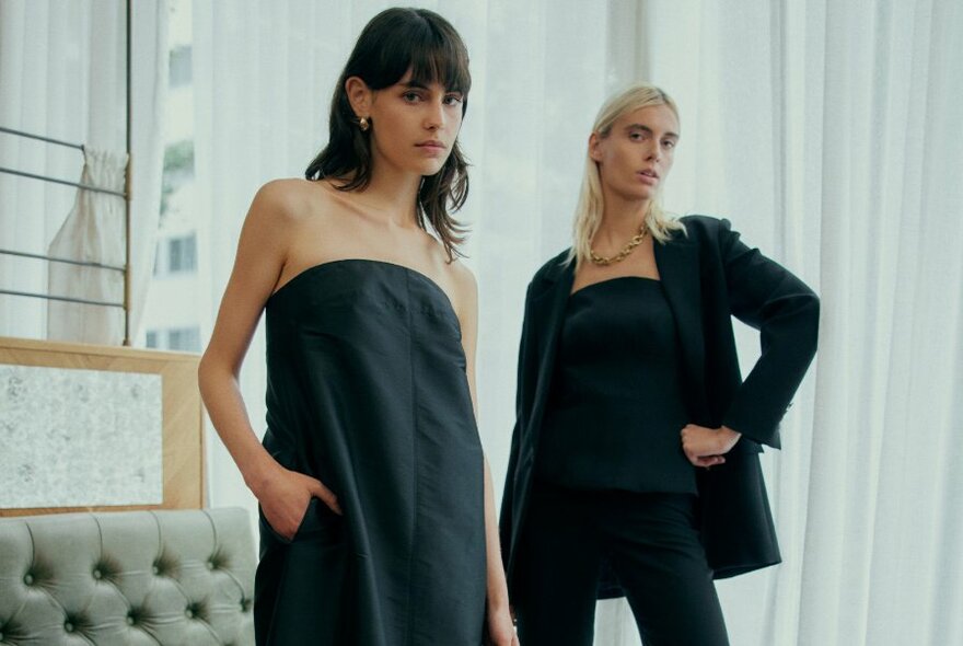 Two women modelling black designer clothes in an interior space, each with a hand on their hips.