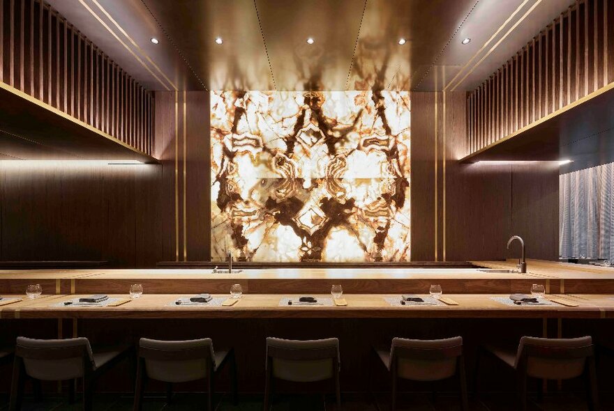 Five high stools facing a bar area that is set for meals, with an illuminated central wall facing the seats.