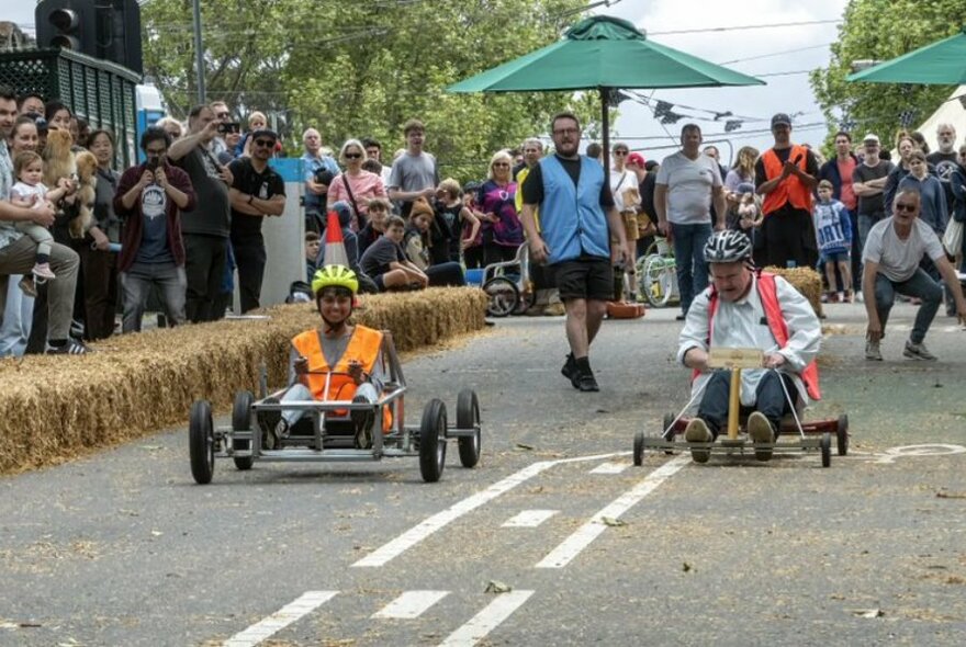 Two people racing unpowered cars on road, hay bales to left; onlookers behind and to left.