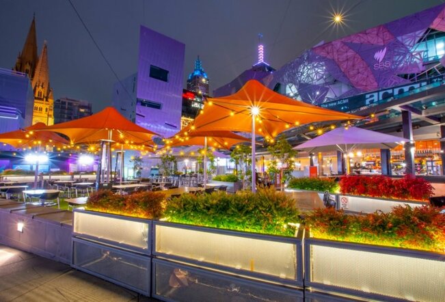A beer garden with orange umbrellas in Federation Square at night.
