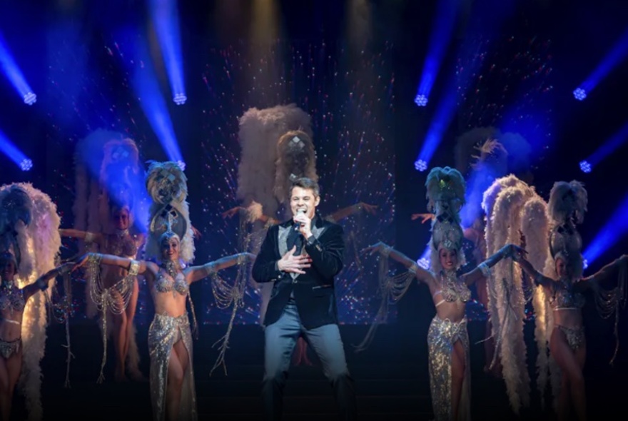 A man singing on stage with showgirls in the background. 