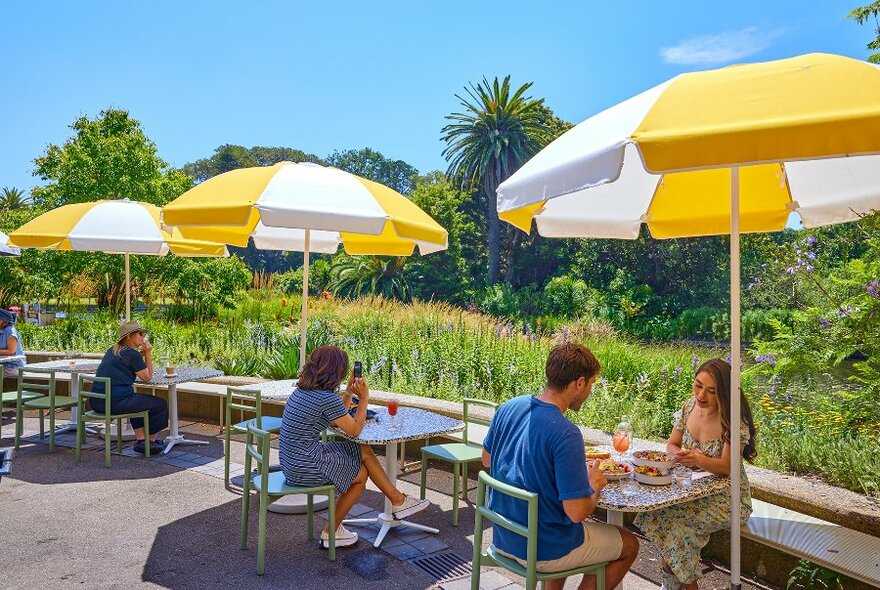 People sitting at small tables under yellow umbrellas near a park. 