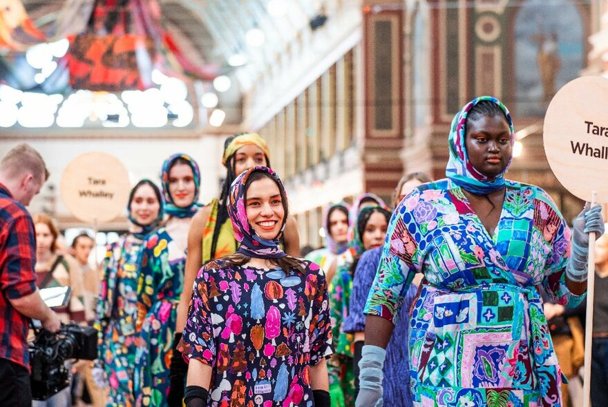 Pop up runway with models walking on it wearing brightly patterned garments, inside a large exhibition hall.