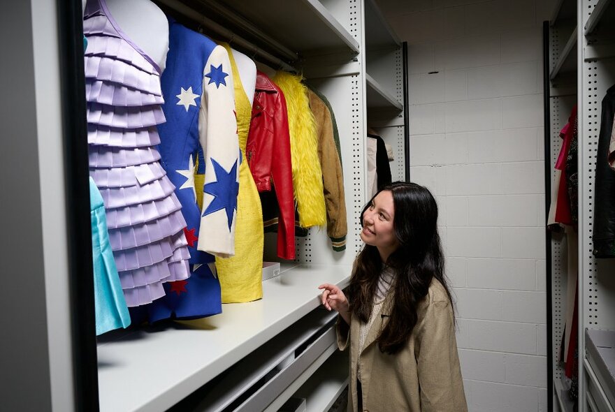 A woman in a walk-in wardrobe space looking at very colourful outfits and smiling. 