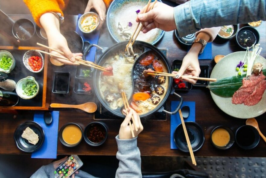 Looking down on a table with a steaming hot pot and four hands holding chopsticks.
