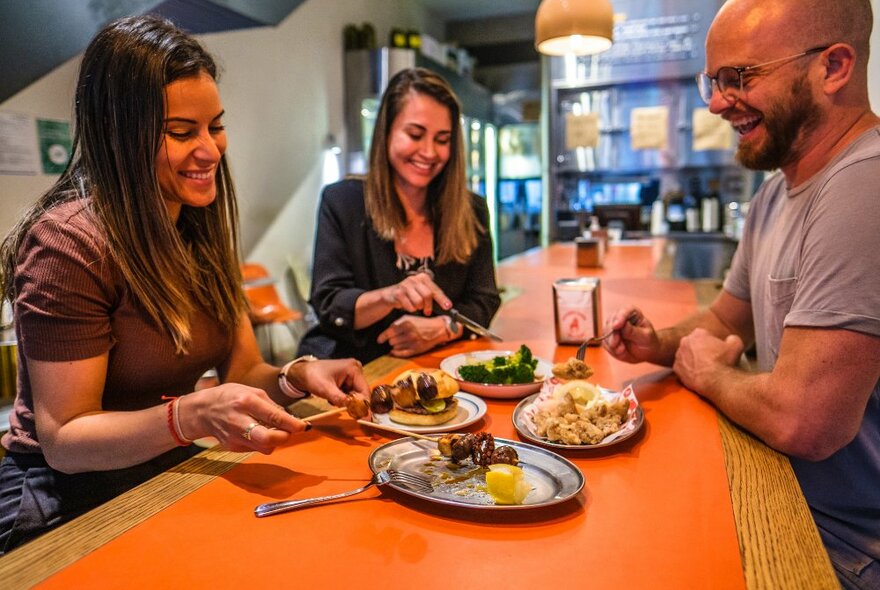 Three people smiling and sharing plates of fried food at a long orange diner table.