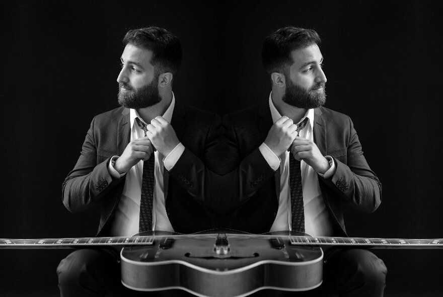 Black and white mirrored image of the guitarist Adam Russo in a suit, straightening his tie, looking to the side, a guitar resting on his lap.