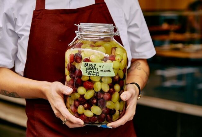Close up shot of a chef in a white shirt, maroon apron holding a big jar of green and red grapes that are fermenting. 