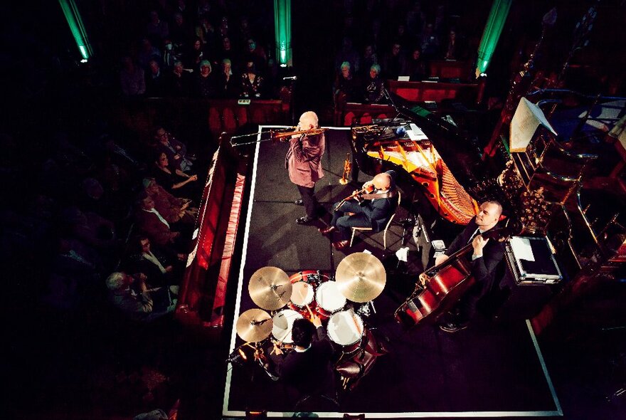 Overhead view of musicians performing on a stage in a darkened venue surrounded by seated onlookers.