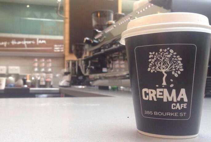Crema Cafe takeaway cup on bench with coffee machine behind the right and counter in far background.