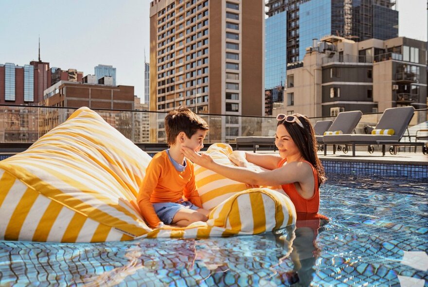 A child sitting on a aqua beanbag, with an adult in the water next to him, enjoying themselves in a hotel's rooftop pool.