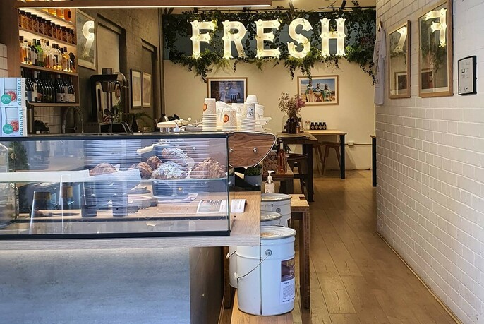 Cartel cafe interior with glass counter, shelves, tables and hanging display featuring the word 'fresh'.
