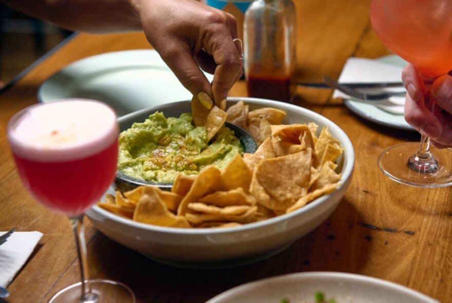 Hand holding corn chip dipping into bowl of guac, next to pink cocktail.