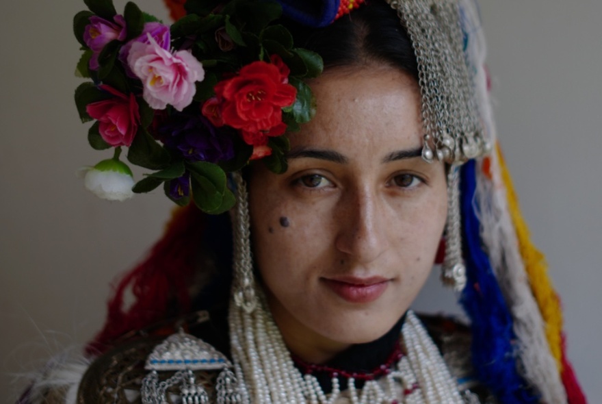 A dark picture of a women from Ladakh in traditional costume with an elaborate headdress containing flowers and fringing. 