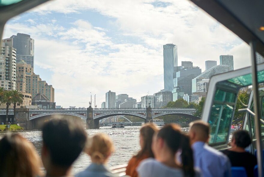 People standing on a boat looking over the river at a view of Princes Bridge and city buildings.