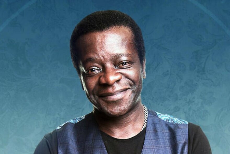 Stephen K Amos smiling, wearing a shiny navy vest over a black top.