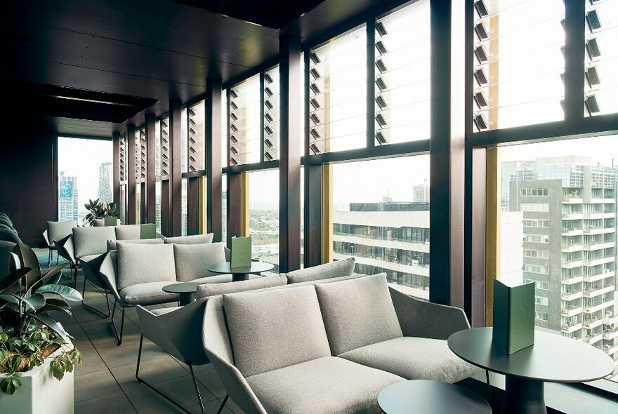 Grey two-seater sofas and small tables arranged next to windows that overlook a city skyscraper view.