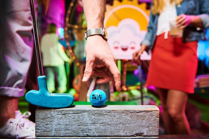 Hand placing a blue golf ball on a putting tee, with golf club nearby and people in the background.