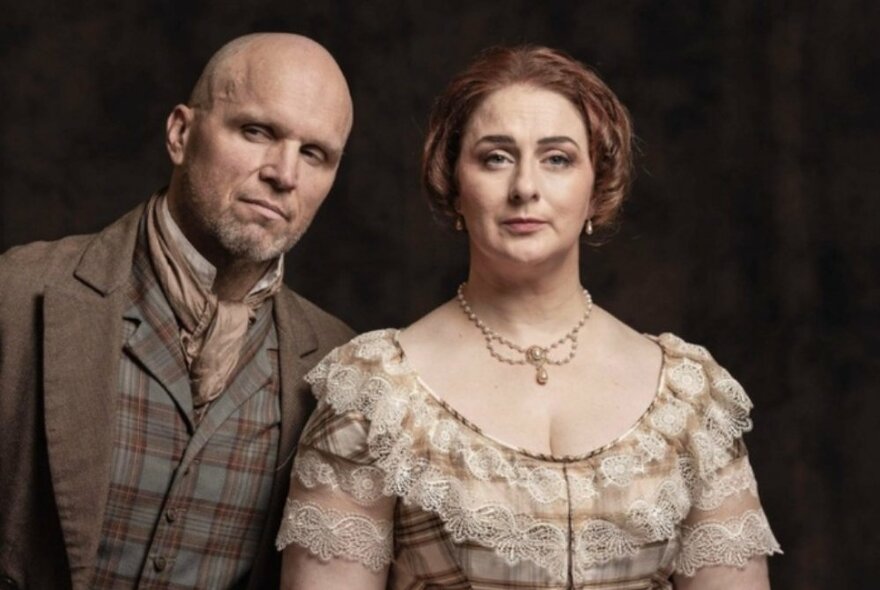 Opera singers in historical character dress, the man wearing a brown waistcoat and cravat, the woman wearing a brown low-necked gown with necklace.