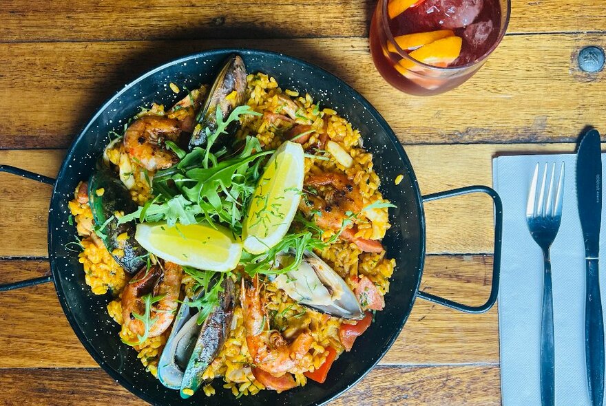 A pan of paella, garnished with lemon wedges, cutlery on a napkin alongside. 