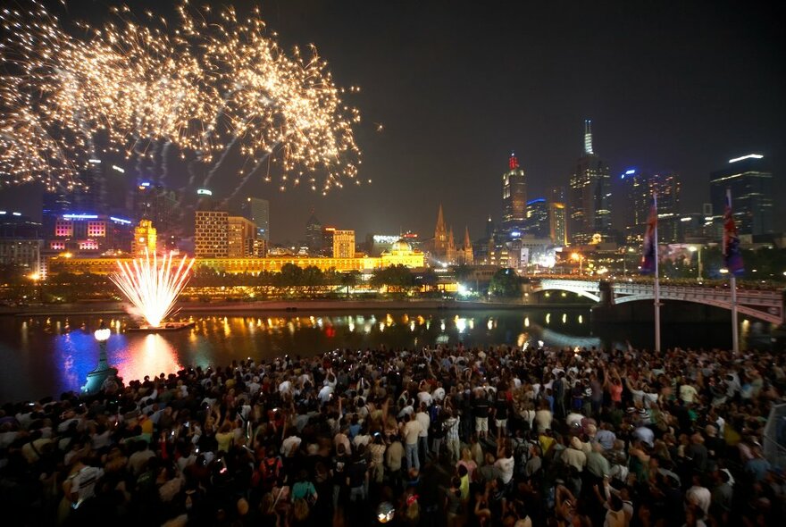 New Year's Eve fireworks exploding over Flinders Street Station and the Yarra River while a crowd of people look on.