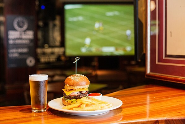 Hamburger and fries on a plate, with a pot of beer on a table, with a television screening sport in the background.