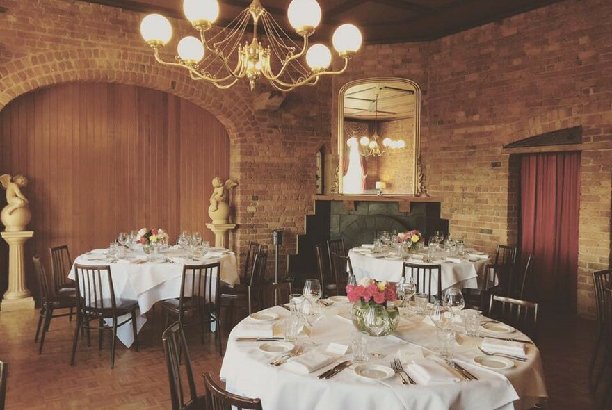 Brick-walled dining room with chandelier, round tables set for dinner with white napery, cherub statues.