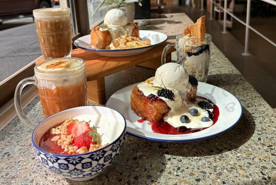 Dishes lined up on a cafe table, including a bowl topped with strawberries, bread with ice cream and mugs of milk drinks.