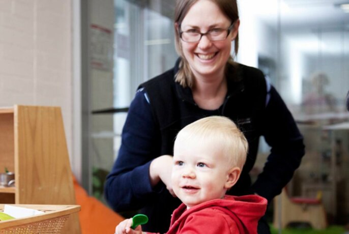 Smiling woman wearing glasses, looking at a toddler in a library setting.