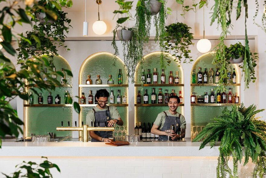 Bartenders preparing drinks behind a white and green bar decorated with plants.