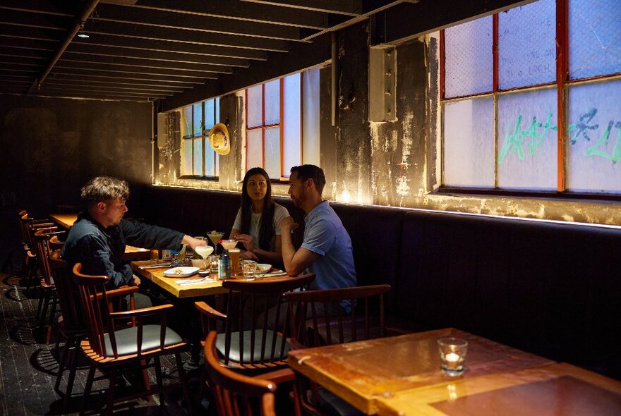 Three people at a table in an empty restaurant with warehouse windows behind, and a delicious array of Mexican food and cocktails on their wooden table.