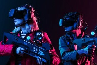 Two figures wearing visual reality goggles and carrying gamer's shooting apparatus in a darkened room.