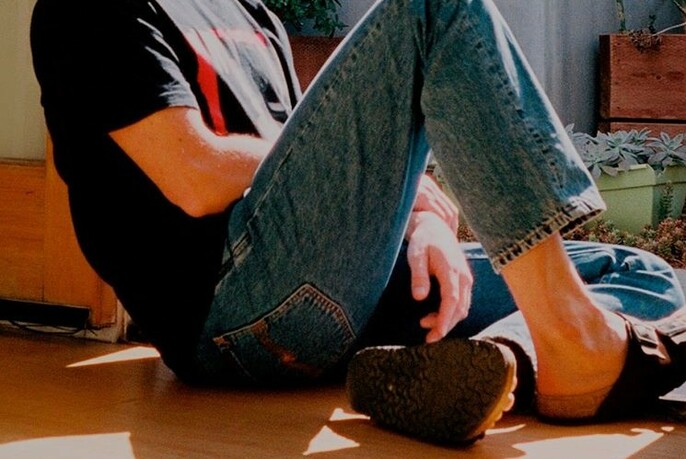 Seated male model wearing Nudie jeans, T-shirt and sandals.
