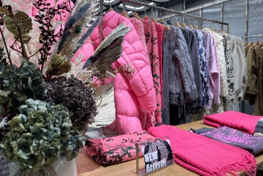 Coats and jackets on a rack, behind table with vase of flowers at front left, and scarves to right.