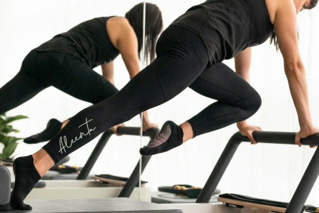 Woman in black active wear resting both arms on a Pilates Reformer machine, with one knee bent forward.