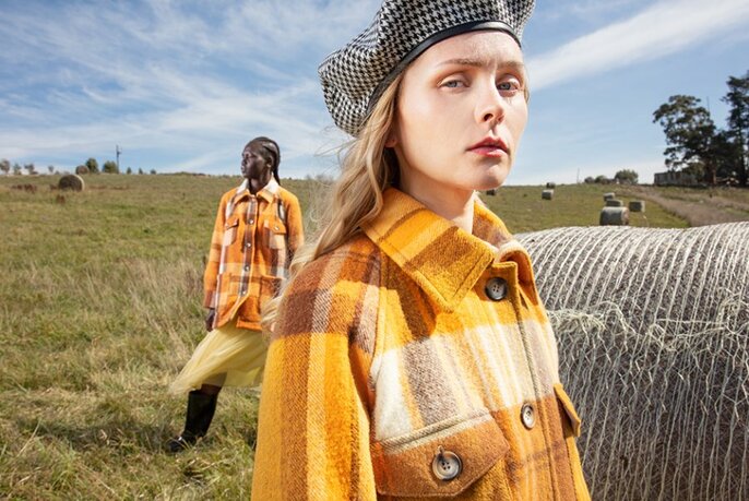 Two models wearing orange and brown coloured coats standing outdoors in a grassy field, with a blue sky visible.