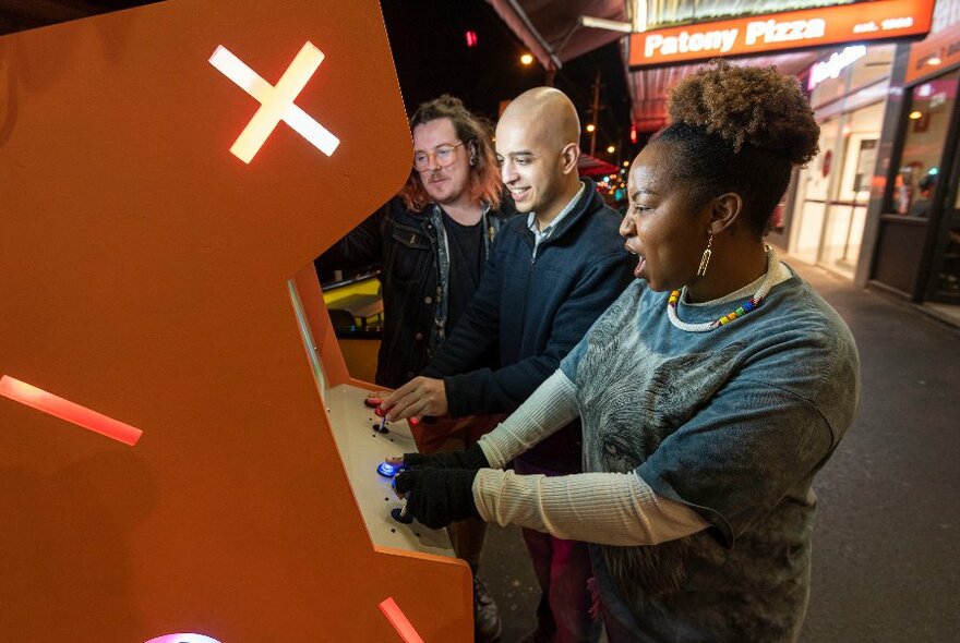 Three adults playing arcade games, the orange machine bearing a large X. 